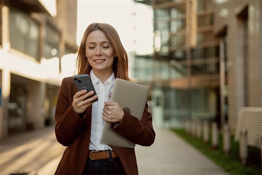Woman looking at phone while holding laptop outside